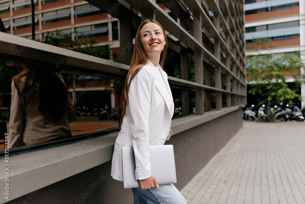 Smiling confident woman with long hair wraith white jacket is holding laptop and posing on background of modern business district in warm sunny day - obrazy, fototapety, plakaty 