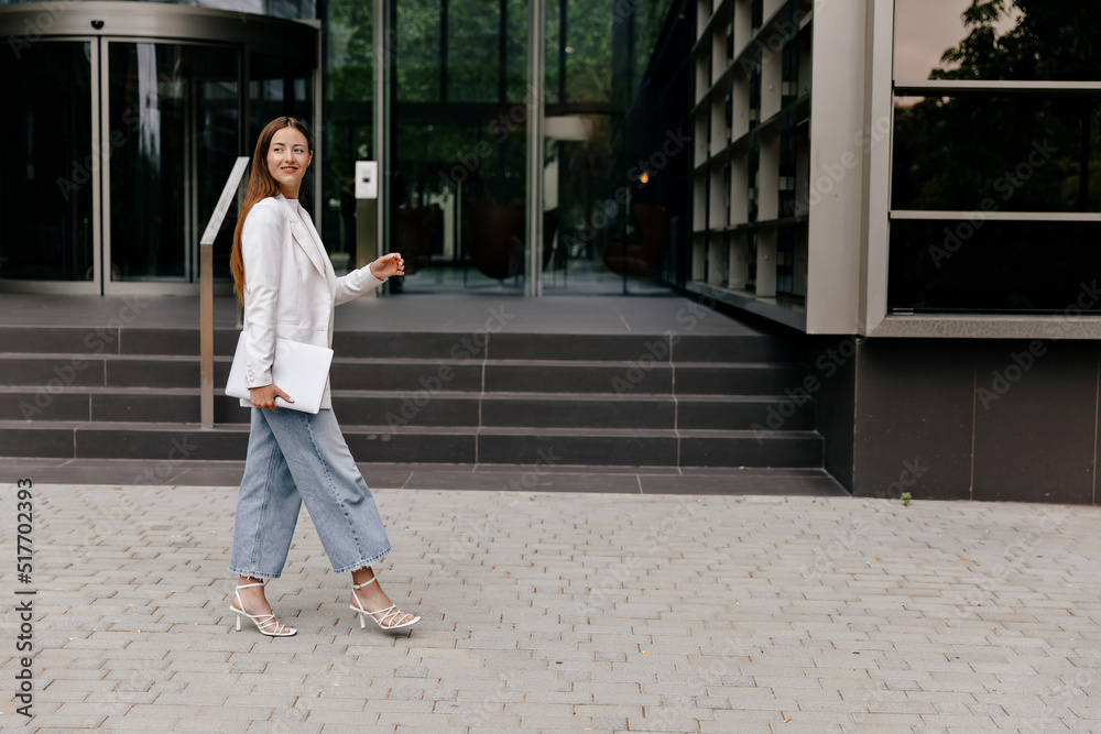 Full-lenght portrait of adorable lovely girl wearing white jacket and jeans and heels walking with laptop to office on background of modern building 