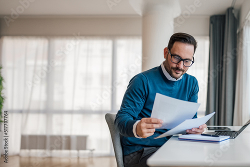 Smiling young executive analyzing business documents. Male professional is working on laptop. He is wearing formals while sitting at office desk.