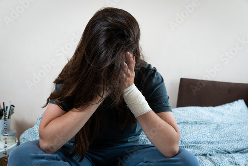 Young unhappy injured woman with a bandage on her hand hides her face. Depressed female scared and stressed suffering from depression and anxiety. Shelter for victims of violence. photo