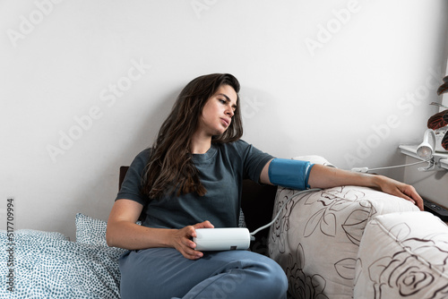 Young sick woman or girl measures her blood pressure with a digital blood pressure monitor. Ill female lying in the bed high blood pressure. photo