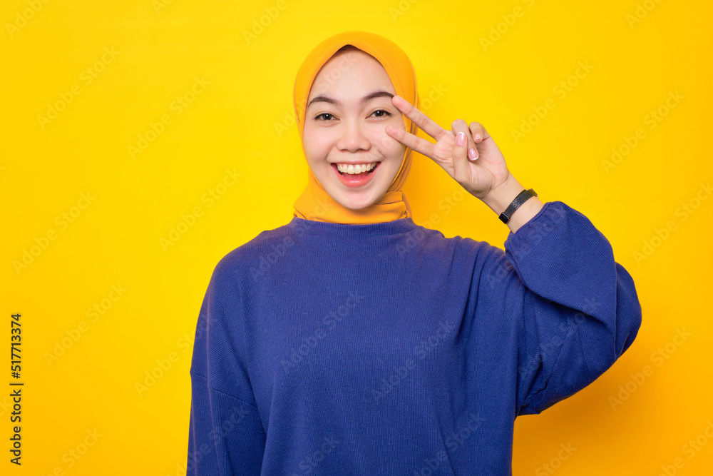 Cheerful young Asian Muslim woman dressed in casual sweater doing peace symbol with fingers over face isolated over yellow background Cheerful young Asian Muslim woman dressed in casual sweater doing peace symbol with fingers over face isolated over yellow background