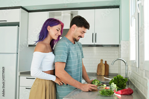 Young woman with colorful hair and her husband cooking food in the open plan kitchen of their new home. Hipster couple spending quality time together. Copy space, interior background.