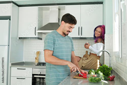 Young woman with colorful hair and her husband cooking food in the open plan kitchen of their new home. Hipster couple spending quality time together. Copy space, interior background.