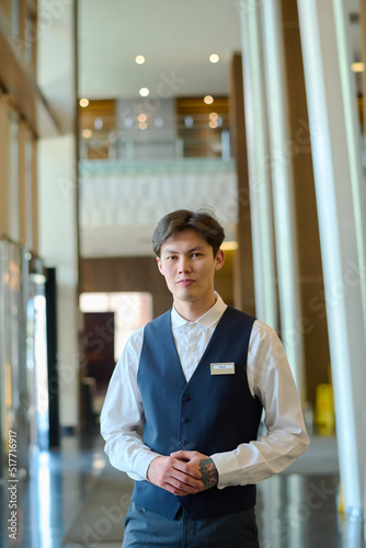 Young manager of hotel or bellboy in uniform standing in front of camera in corridor or lounge and waiting for new guests photo