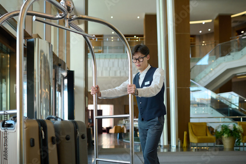 Young bellboy in uniform pushing cart with suitcases of hotel clients or guests while moving along corridor or crossing lounge photo