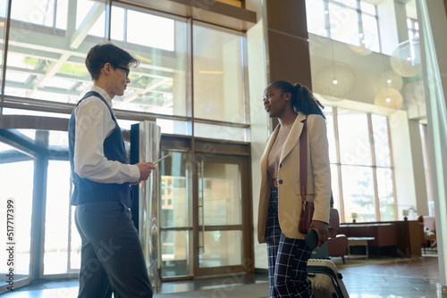 Young bellboy in uniform meeting black woman in smart casualwear pulling suitcase in lounge of large modern luxurious hotel
