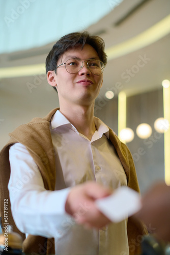 Asian guy in eyeglasses and white shirt passing keycard to receptionist by counter in lounge while going to leave hotel for work