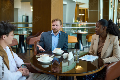 Two young businessmen and black woman in formalwear discussing points of new project while sitting by table in hotel lounge