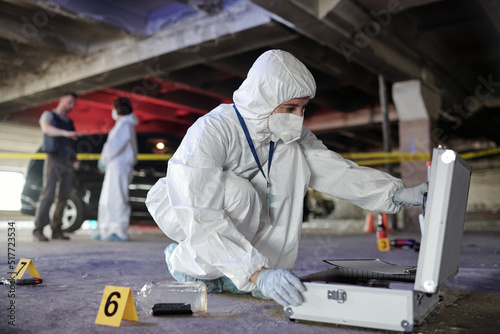 Young female criminological expert in coveralls opening briefcase with working supplies during inspection of evidences on crime scene