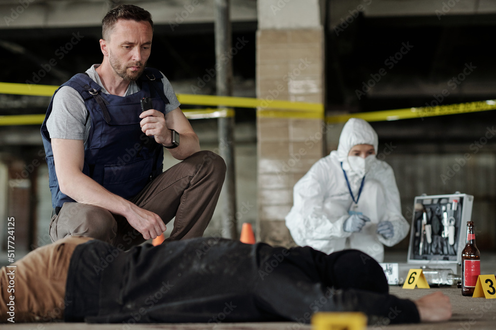 Young serious policeman in bulletproof vest squatting by dead body while inspecting crime scene surrounded by yellow tape