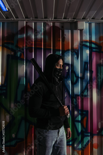 Side view of african american hooligan in mask and hoodie holding baseball bat near graffiti on wall