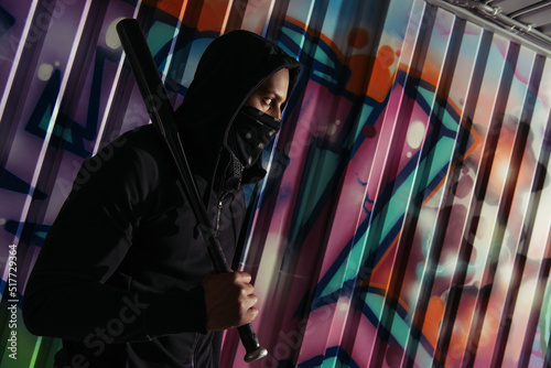 Side view of african american bandit in mask holding baseball bat near graffiti on wall