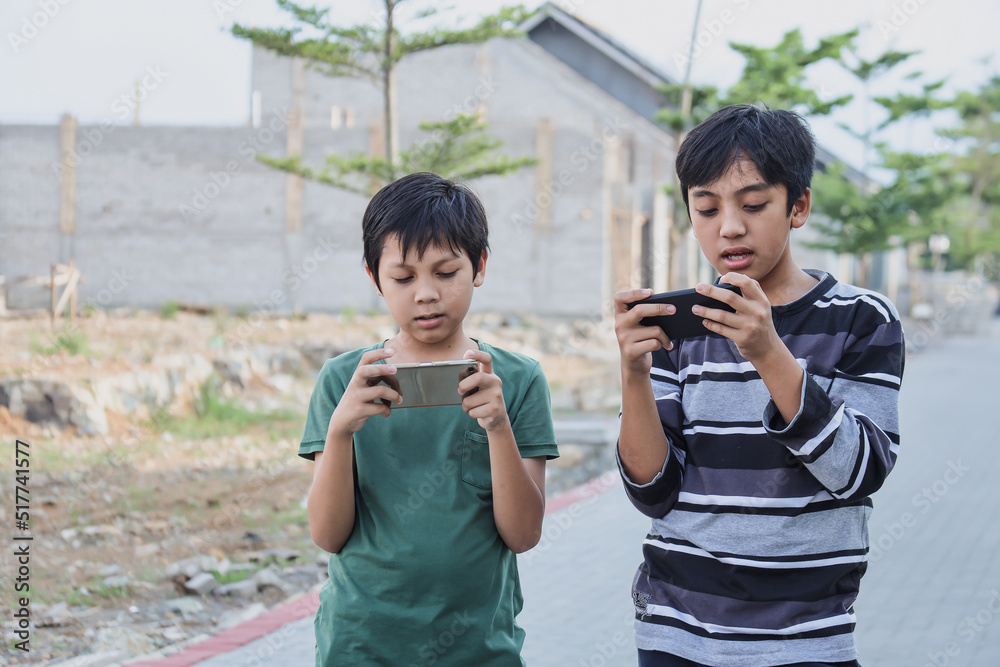 Two kids with gadgets. Brothers surfing the net or playing online games on smartphone and digital tablet outside while standing. Modern communication and gadget addiction concept. Two kids with gadgets. Brothers surfing the net or playing online games on smartphone and digital tablet outside while standing. Modern communication and gadget addiction concept.