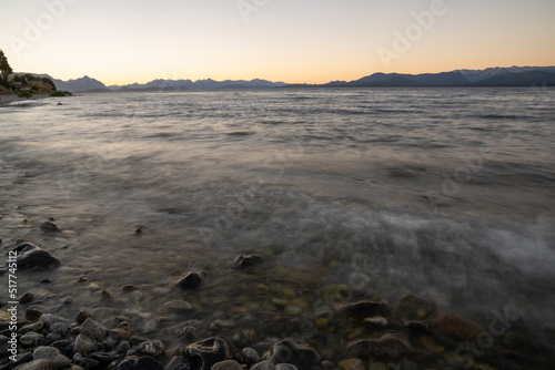 Long exposure shot of Nahuel Huapi lake at sunset. Beautiful blurred water effect, the rocky shore and Andes mountains in the horizon with a magical dusk light.