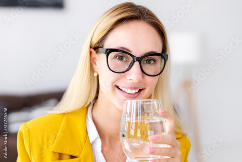 Portrait of beautiful blonde businesswoman with glass of water. Young woman with eye glasses drinking clean water, looking at the camera and smiling. Recuperation, healthy lifestyle concept