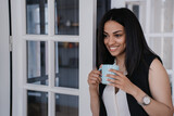 Grateful toothy smiling afro american young woman having break holding cup of coffee smiling leaning on door at office. Cheerful Brazilian businesswoman relaxing at home, business and education.
