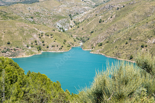 Breath-taking view from the top on green lake surrounded by picturesque mountain alley with trees and bushes in sunny summer day with green trees and bushes in the foreground.