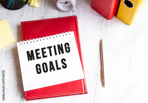 Notepad with the text MEETING GOALS on a wooden table. Red diary and pen.