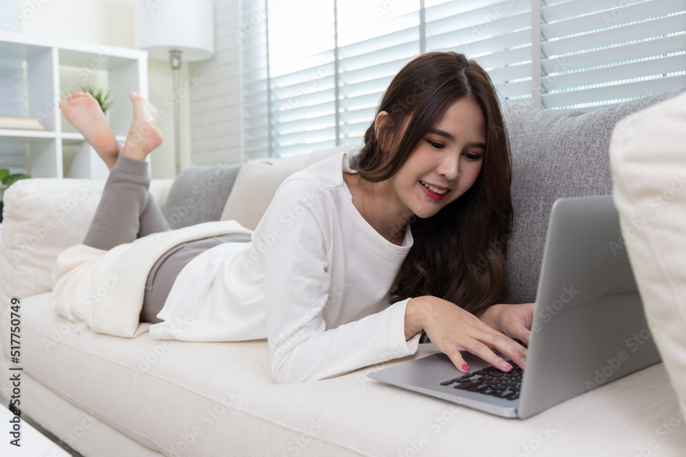 Young Asian woman using laptop computer checking social media application on website online and writing blog and lying on sofa in living room at home Young Asian woman using laptop computer checking social media application on website online and writing blog and lying on sofa in living room at home