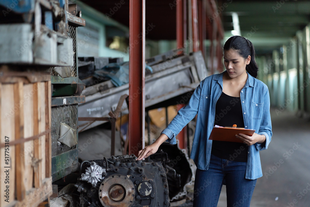 Asian woman worker working at old automotive spare parts warehouse. Woman warehouse worker checking old engine, motor, machine at the garage industry factory.