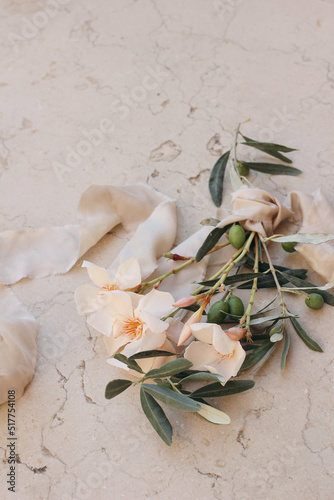 Summer wedding floral composition. Closeup of apricot oleander blossom, flowers and green olive tree branches. Beige marble background with silk ribbon. Blurred background, selective focus. Vertical.
