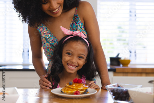 Midsection of happy biracial mother and daughter with tempting pancakes on table at home