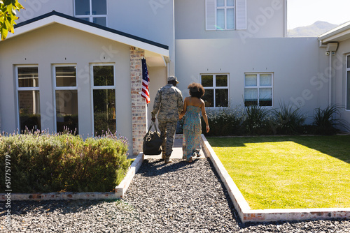 Rear view of multiracial army soldier in camouflage holding wife's hand and going inside house