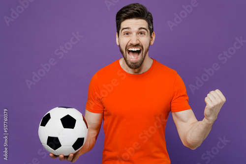 Young fun excited happy fan man he 20s wearing orange t-shirt cheer up support football sport team hold in hand soccer ball watch tv live stream do winner gesture isolated on plain purple background.