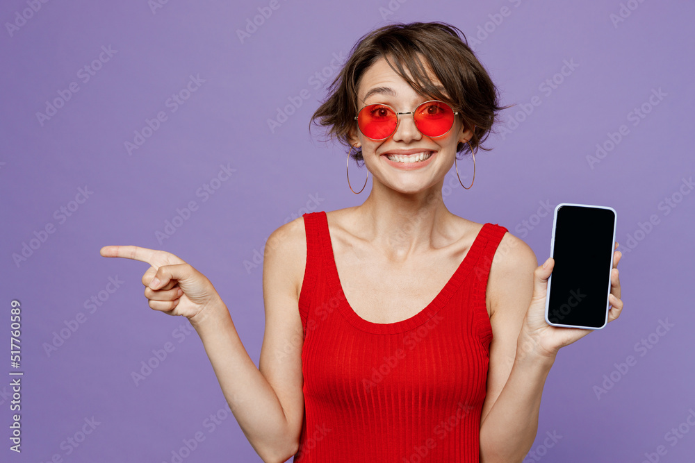 Young amazed excited happy woman 20s she wear red tank shirt eyeglasses hold in hand use mobile cell phone with blank screen workspace area point index fingre aside isolated on plain purple backround
