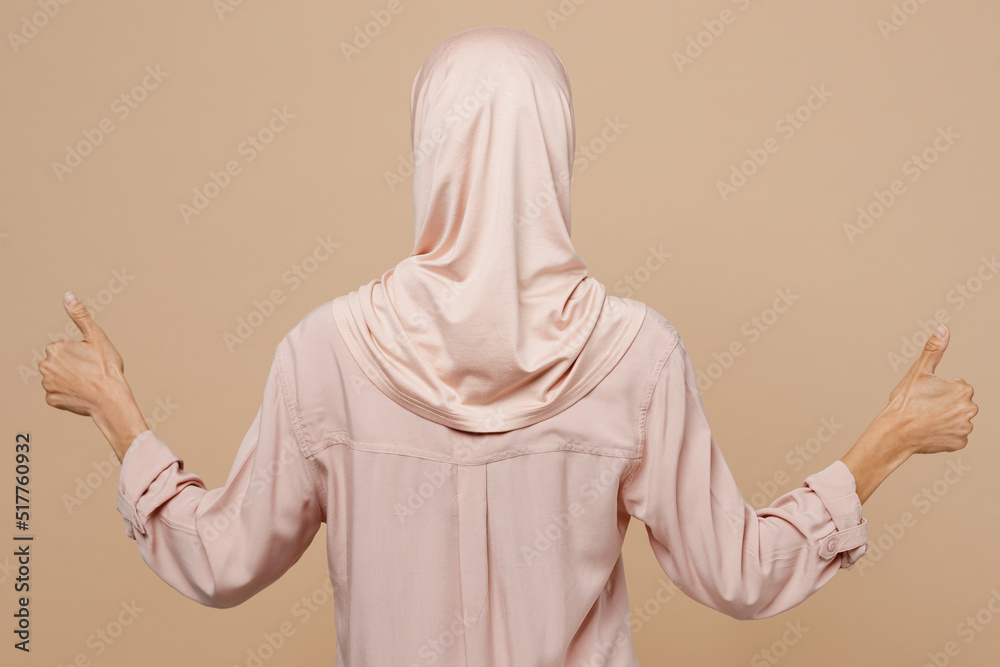 Fototapeta premium Back rear view young arabian asian muslim woman she wearing abaya hijab pink clothes show thumb up isolated on plain pastel light beige background. People uae middle eastern islam religious concept.