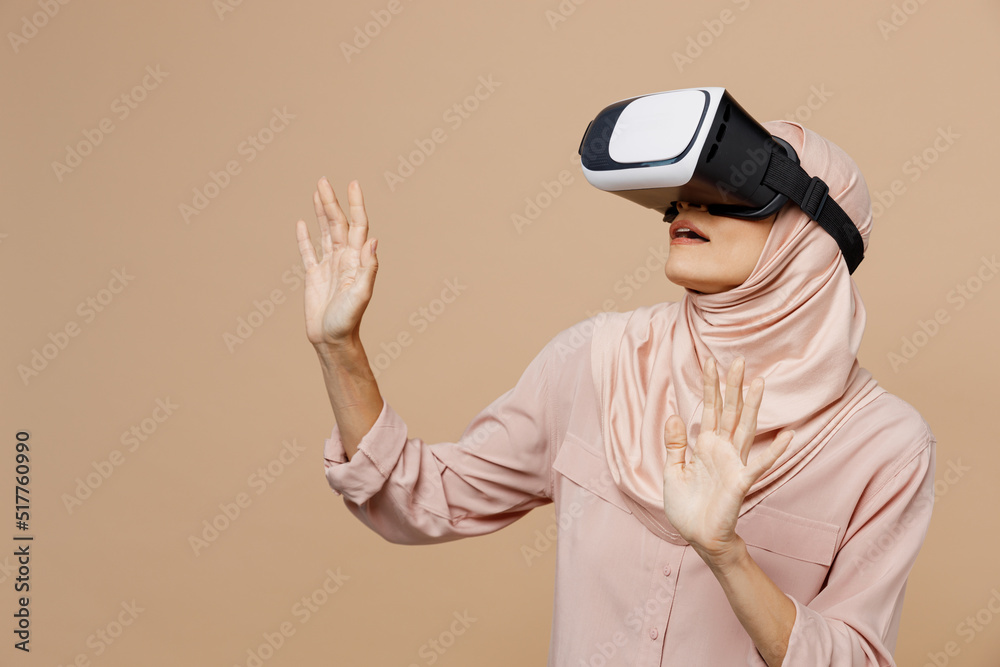 Young arabian asian muslim woman she wear abaya hijab pink clothes watching in vr headset pc gadget isolated on plain pastel light beige background. People uae middle eastern islam religious concept.