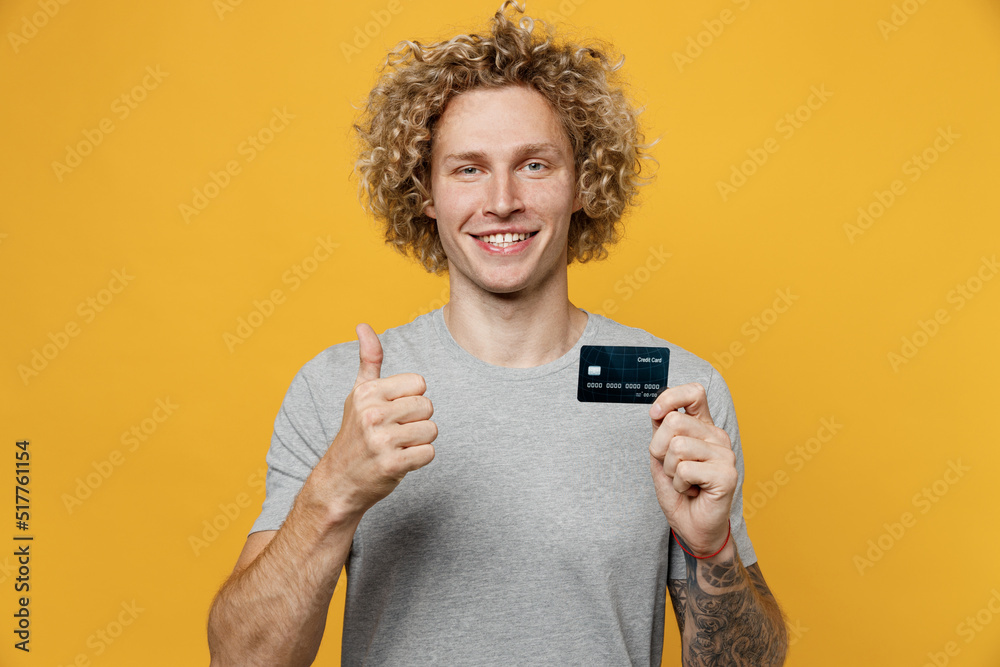 Young smiling cheerful fun cool caucasian man 20s he wear grey t-shirt look camera hold in hand mock up of credit bank card show thumb up isolated on plain yellow backround. People lifestyle concept. Young smiling cheerful fun cool caucasian man 20s he wear grey t-shirt look camera hold in hand mock up of credit bank card show thumb up isolated on plain yellow backround. People lifestyle concept.