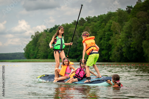 Children having fun on SUP board waving it in water of lake and laughing on cloudy summer day outdoors