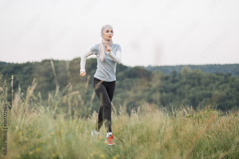Happy arabian woman in hijab in activewear running at summer park. Young woman spending free time activity on fresh air. Smiling muslim female jogging during outdoors workout. Happy arabian woman in hijab in activewear running at summer park. Young woman spending free time activity on fresh air. Smiling muslim female jogging during outdoors workout.