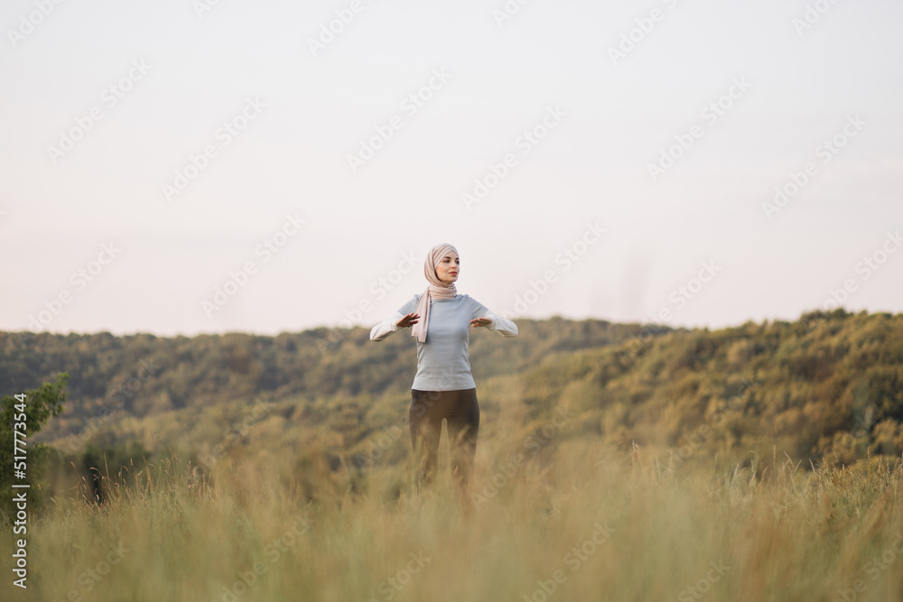 Full length portrait of muslim smiling woman dressed in religious beige hijab stretching her hands while doing workout in green park outdoors. Copy space.