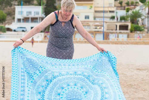 Short haired woman with glasses and wearing printed dress spreads blue and white towel on the beach.