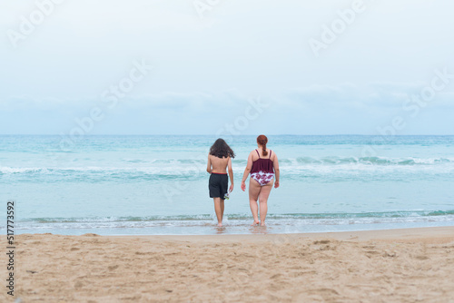 Boy with long hair and bermuda shorts and a young adult entering the sea for a swim. There is copy space.