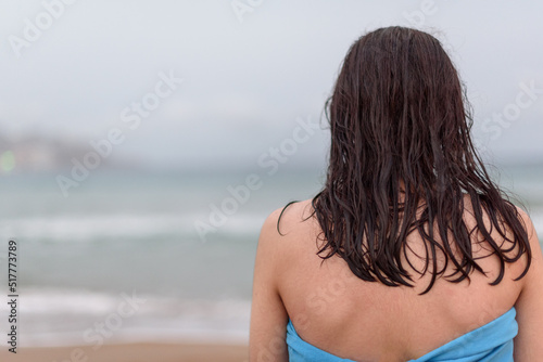 Teenager with long hair looking towards the horizon on the beach after bathing, while drying himself wrapped in a blue towel.