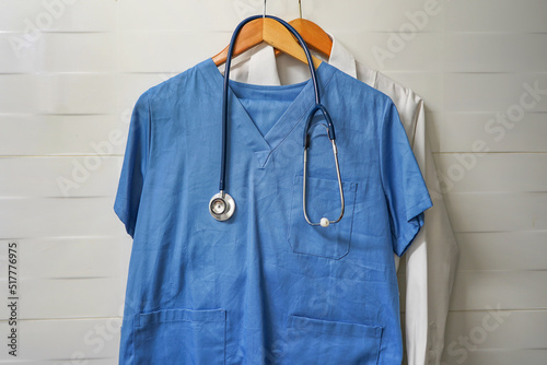 Closeup doctors scrus with stethoscope. Blue surgical smock with stethoscope on wooden hanger on ceramics wall background. Closeup of doctor's scrubs with stethoscope and lab coat on hangers photo