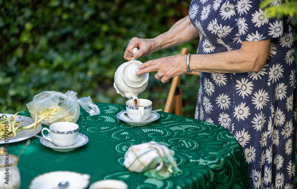 Fototapeta premium old woman in dress pours coffee into cup, natural table in summer garden, everyday life, drinking coffee