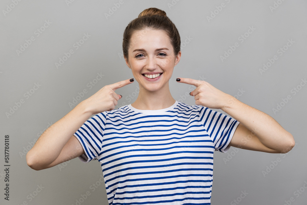 Portrait of pretty cheerful woman wearing striped T-shirt with bun hairstyle points index fingers at smile shows white teeth, looking at camera. Indoor studio shot isolated on gray background. Portrait of pretty cheerful woman wearing striped T-shirt with bun hairstyle points index fingers at smile shows white teeth, looking at camera. Indoor studio shot isolated on gray background.