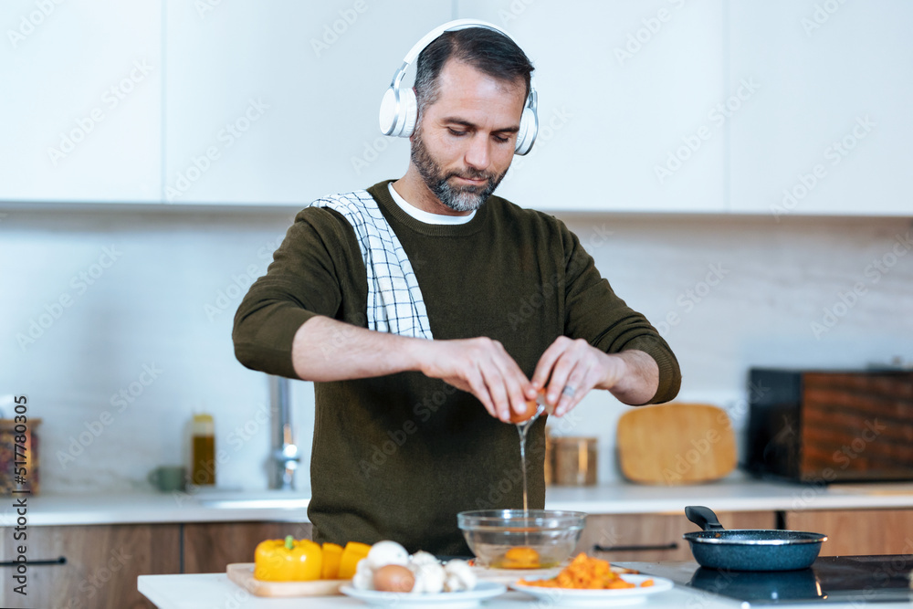 Handsome mature man preparing an omelette while listening music with headphones in the kitchen at home.