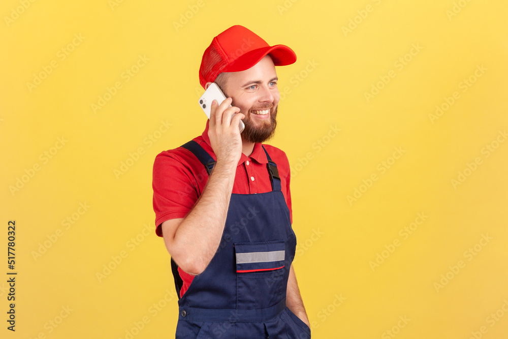 Portrait of happy courier or repairman in overalls taking phone with client, specify the time and place of completing work or delivery of goods. Indoor studio shot isolated on yellow background. Portrait of happy courier or repairman in overalls taking phone with client, specify the time and place of completing work or delivery of goods. Indoor studio shot isolated on yellow background.