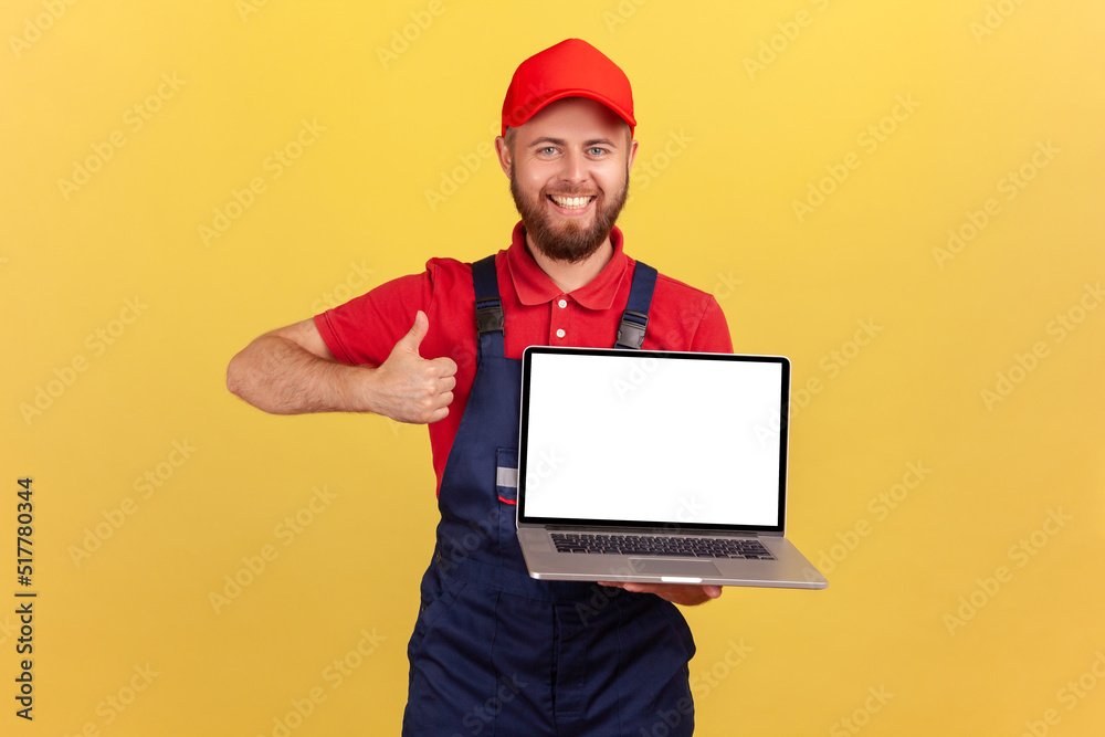 Portrait of satisfied happy worker man in uniform standing and holding laptop with blank screen, advertisement area, showing thumb up. Indoor studio shot isolated on yellow background. Portrait of satisfied happy worker man in uniform standing and holding laptop with blank screen, advertisement area, showing thumb up. Indoor studio shot isolated on yellow background.