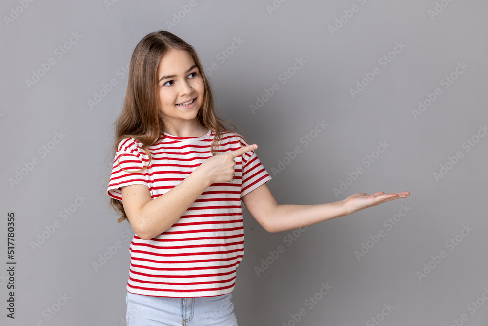 Portrait of little girl wearing striped T-shirt presenting advertising area on her palm and pointing to copy space, holding empty place for commercial. Indoor studio shot isolated on gray background. Portrait of little girl wearing striped T-shirt presenting advertising area on her palm and pointing to copy space, holding empty place for commercial. Indoor studio shot isolated on gray background.