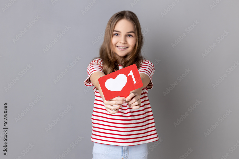 Portrait of smiling little girl wearing striped T-shirt showing heart like icon of social media and looking at camera with cute smile. Indoor studio shot isolated on gray background. Portrait of smiling little girl wearing striped T-shirt showing heart like icon of social media and looking at camera with cute smile. Indoor studio shot isolated on gray background.