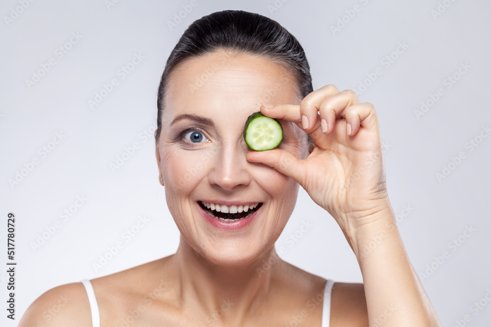 Skin care natural beauty and cosmetology concept. Closeup of excited middle aged woman covering eye with slice of fresh cucumber, makes mask for face. Indoor studio shot isolated on gray background. Skin care natural beauty and cosmetology concept. Closeup of excited middle aged woman covering eye with slice of fresh cucumber, makes mask for face. Indoor studio shot isolated on gray background.