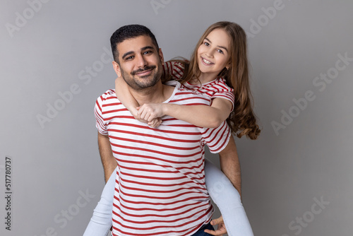 Portrait of positive optimistic father and daughter in striped T-shirts spending time together, dad holding his charming little kid on his back. Indoor studio shot isolated on gray background.