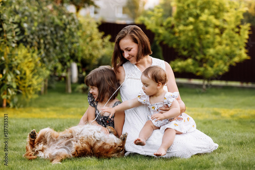 Family day, mother's day. Beautiful smiling young mom and two child daughters cuddling happy domestic dog on the backyard lawn. Idyllic family having fun with pet outdoors on summer holiday.